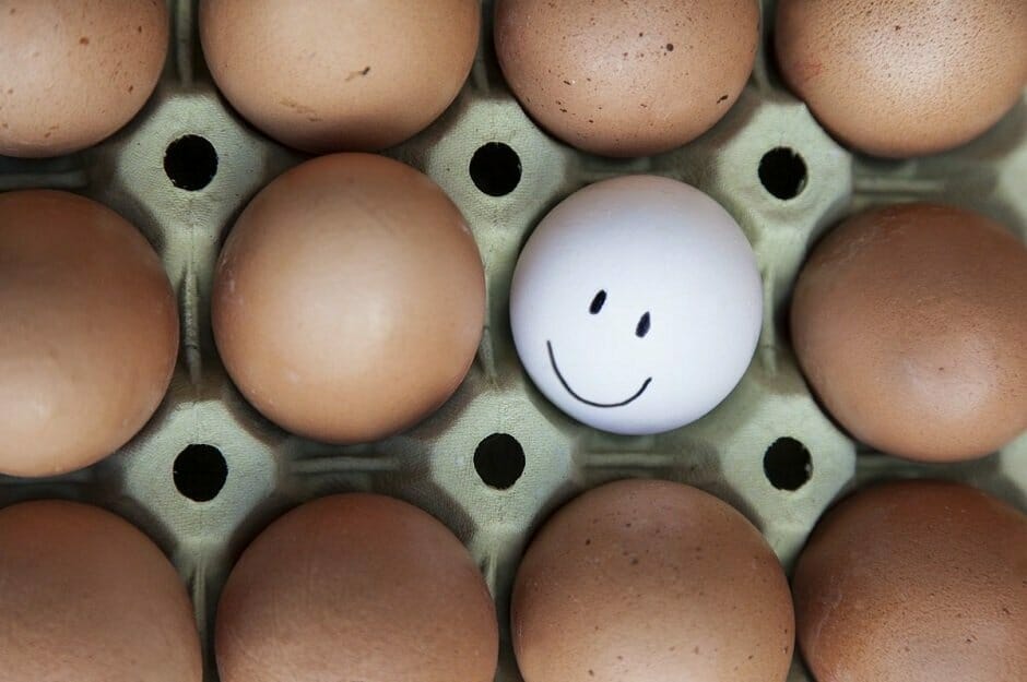One white egg with drawn on smiley face in carton among brown eggs.